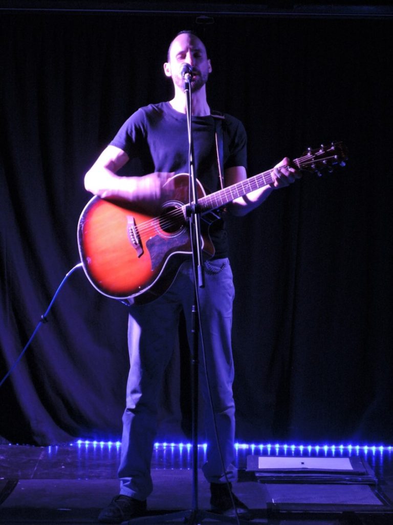 Catching the Ghost - A profile shot of a tall white man with short dark hair and a goatee stands on a dimly lit stage. He sings into a microphone, placed on a stand and strums of a guitar.