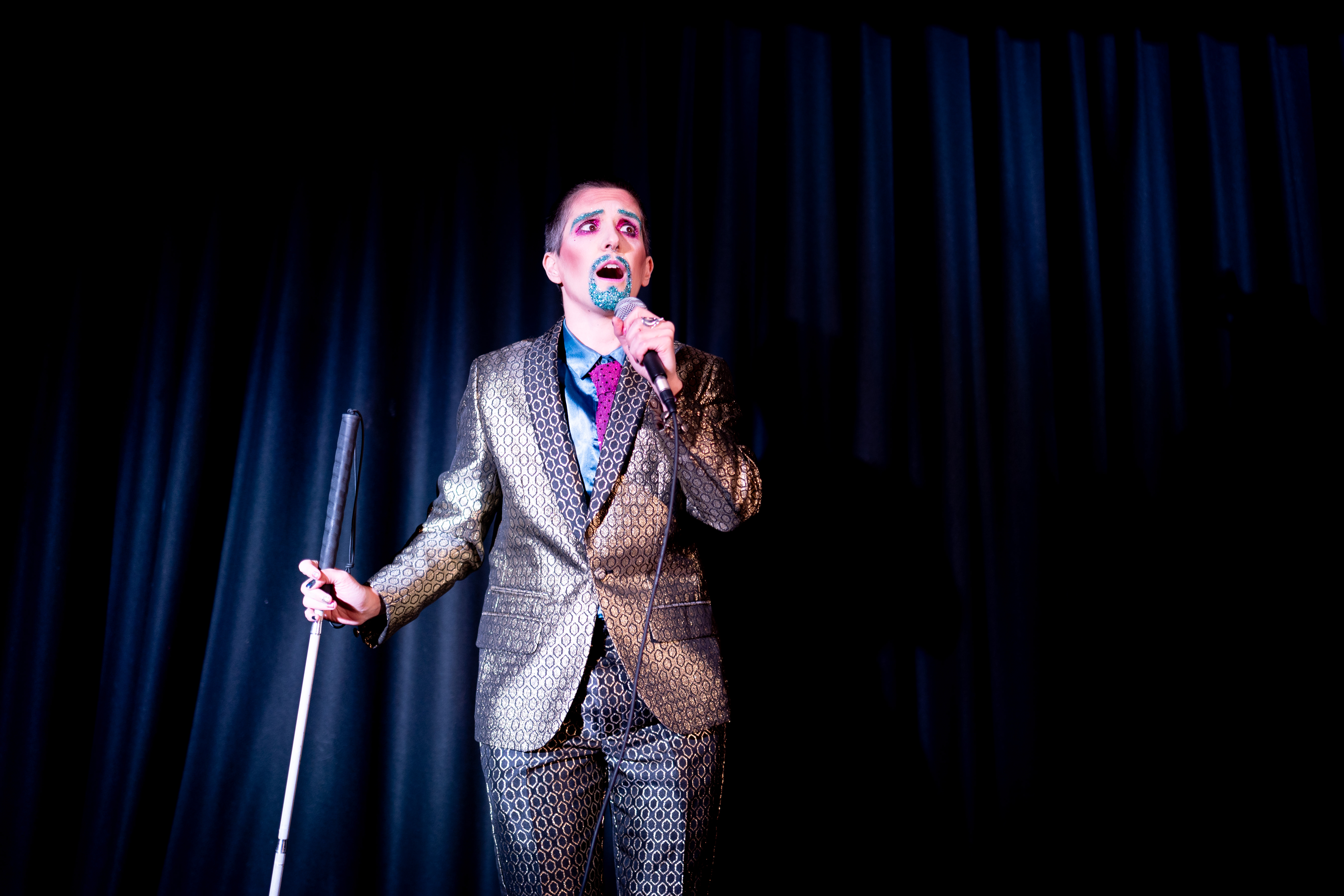 A white drag king wearing a shiny bronze suit, eyes wide, and talking on a microphone