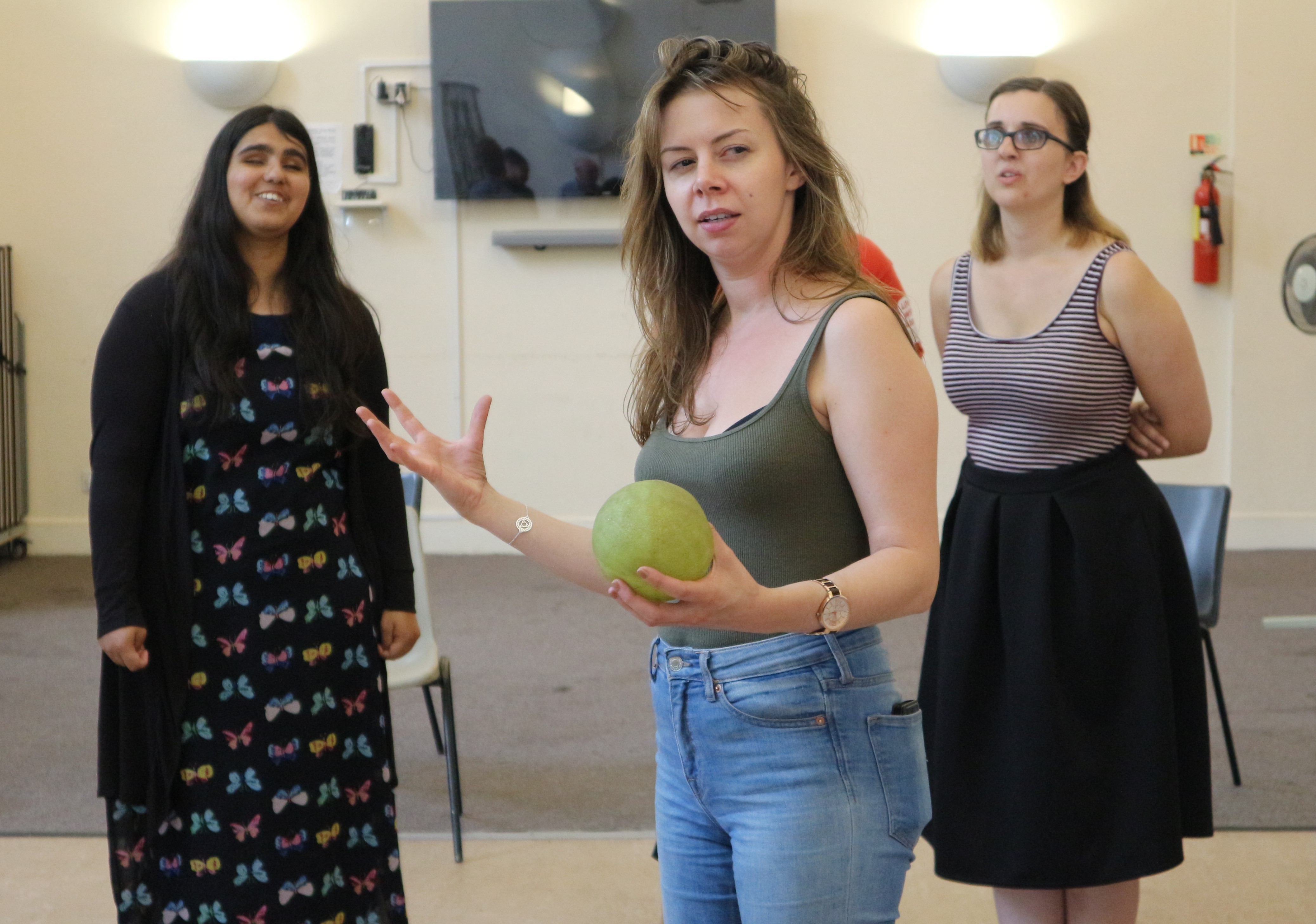 A white woman stands in front of two workshop participants. The woman has a light green ball in her hand, looking thoughtfully across the room while gesticulating