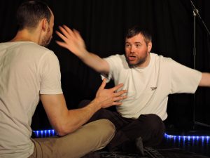 Two white men sit on the floor opposite one another. The man facing the camera is stretching his arm out toward the other man. 