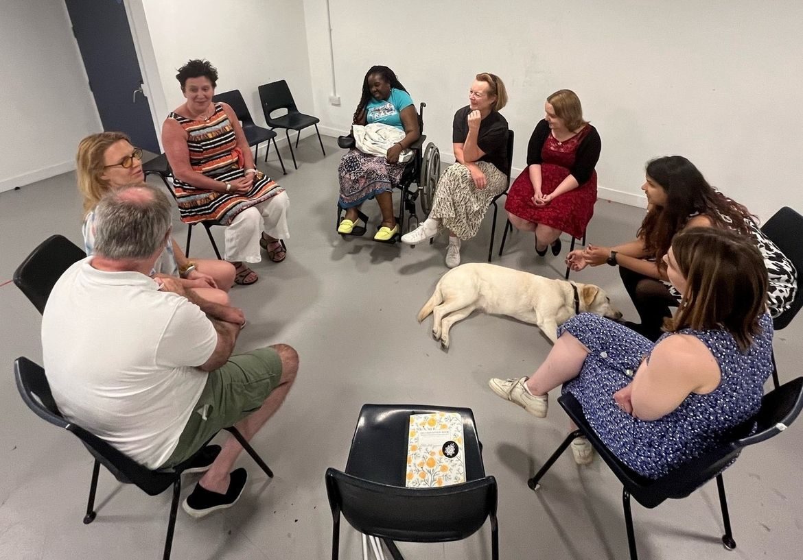 Eight No Dramas attendees sit in chairs in a circle, talking and smiling in the basement of The Camden People’s Theatre. They are a diverse group of people, wearing casual clothing. There is one man and seven women with one attendee in a wheelchair. A guide dog lies on the floor in the middle of the circle. The room is bare and brightly lit with a grey floor and white walls. 