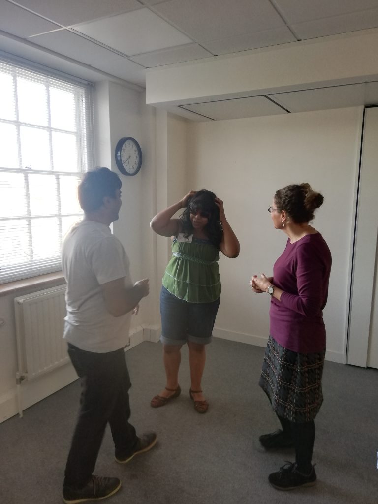 A black woman, stands centrally facing an Asian man and white woman in small triangle. The black woman has her hands on top of her head and the others step towards her. 