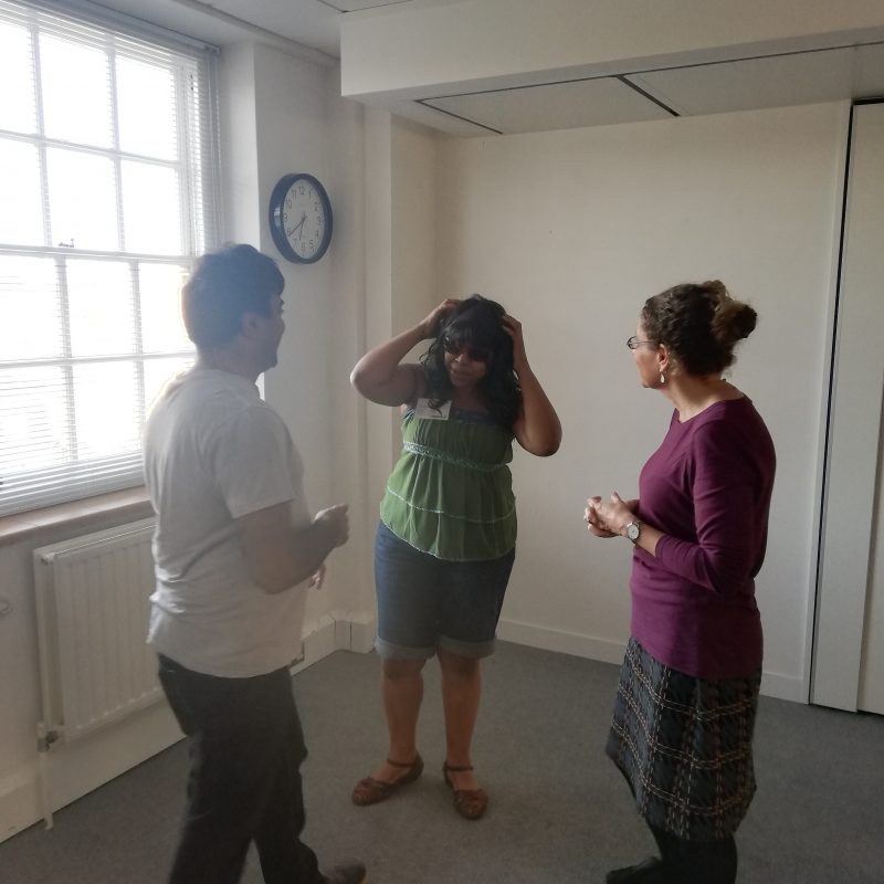 Three woman standing in a circle facing one another. The woman in the centre has her hands drawn to her head, while the woman on the left takes a step towards her.