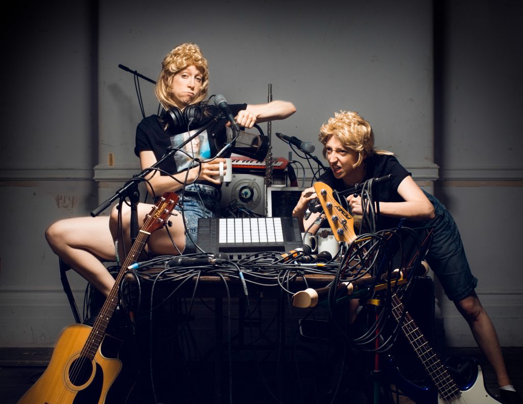 Two white women in blond curly hair pieces perch on a table full of cables entwined with musical instruments and microphones