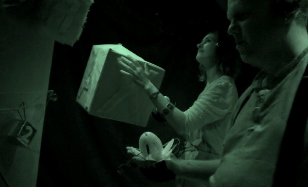 Facing side on to the camera, a woman clutches a large cardbox box. Her face is tilted. A man in the foreground holds a lotus in his hands.