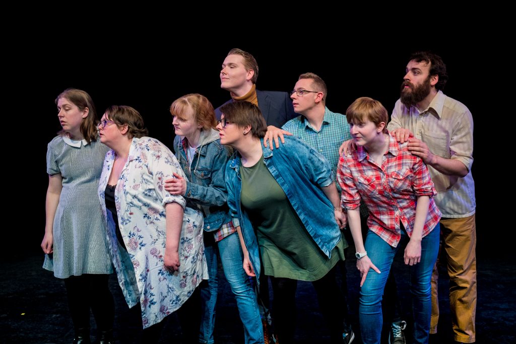 A mixed group of male and female actors stand on stage in a cluster. They are all looking off to their right, leaning in that direction.