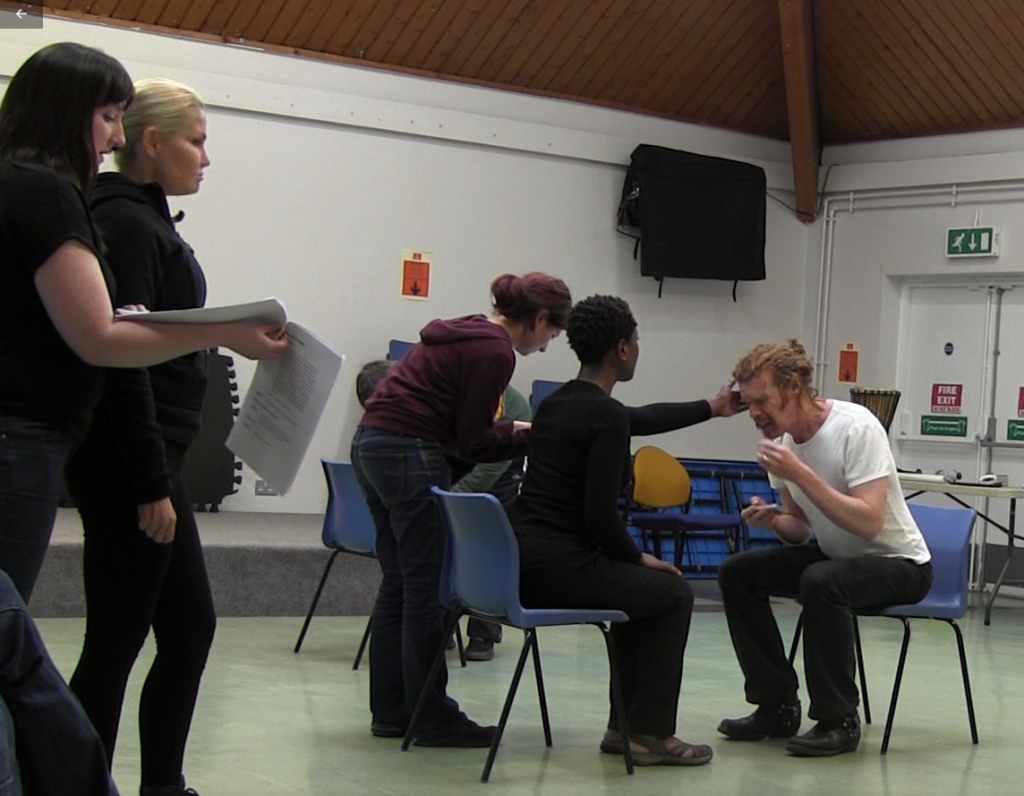 A white man with tied back ginger hair sits on a chair leaning towards a black woman who is reaching out her arm and touching his face. Behind them is a white man with short dark hair who is making noises with a keyboard, and metal cups. To the left of the image stand two white women, the one with dark hair is whispering in the ear of the one with light blonde hair.