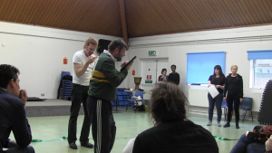 Two white male performers in a rehearsal room reading from their script with audience members watching from the foreground.