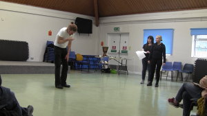 A white man stands hunched reading from his phone. To the side, two women stand close together. The audience watches from the foreground.