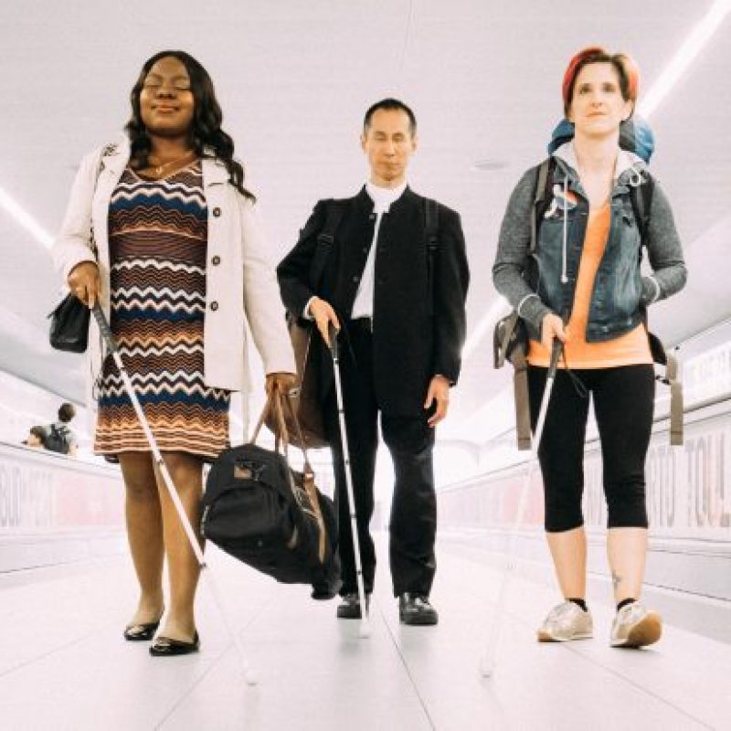 A long corridor framed by two walking escalators typically found in airports. From left to right three visually impaired people striding towards us - a smiling black woman carrying an overnight bag, an East Asian man wearing a suit, and a young white woman with a backpack strung over her shoulders.