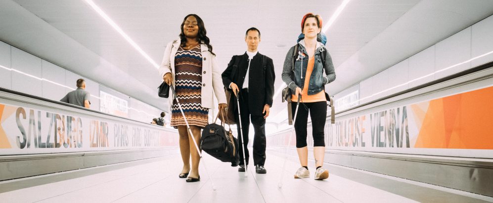 A long corridor framed by two walking escalators typically found in airports. From left to right three visually impaired people striding towards us - a smiling black woman carrying an overnight bag, an East Asian man wearing a suit, and a young white woman with a backpack strung over her shoulders.