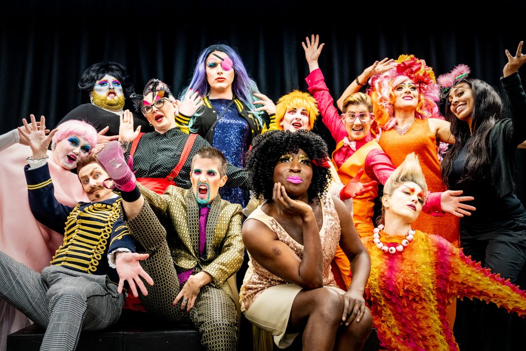 A celebratory group shot of visually impaired drag artists, all dressed in a variety of colourful and outlandish costumes. Some are posing while others grin and have their hands in the air.