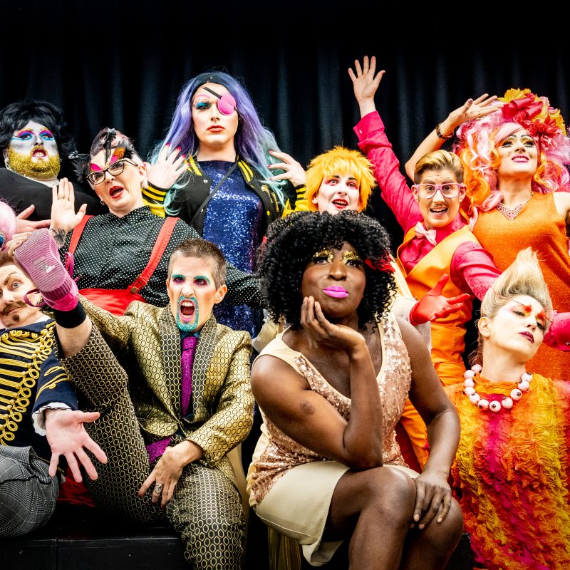 A celebratory group shot of visually impaired drag artists, all dressed in a variety of colourful and outlandish costumes. Some are posing while others grin and have their hands in the air.