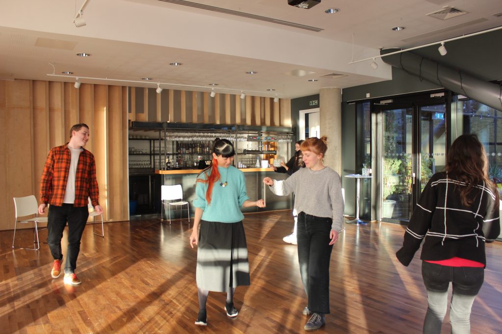 Two young women - an East Asian and white woman walking side by side with a chopstick balanced vertically from each of their fingertips. Three other workshop participants are walking around the room.