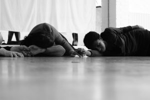 Two young women lying on their sides on the floor, in a position you would use for resting or sleeping. Their eyes are closed and they are holding hands. Black and white image.