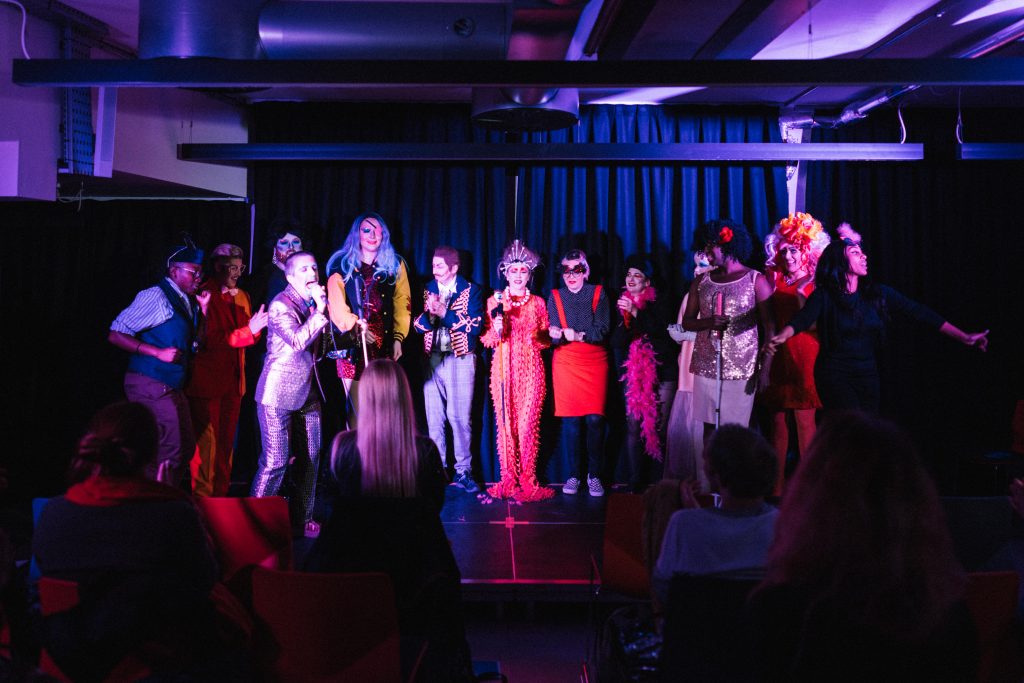The drag performers standing side by side on the stage, some of them clapping. A white drag king stands on the left singing into the microphone