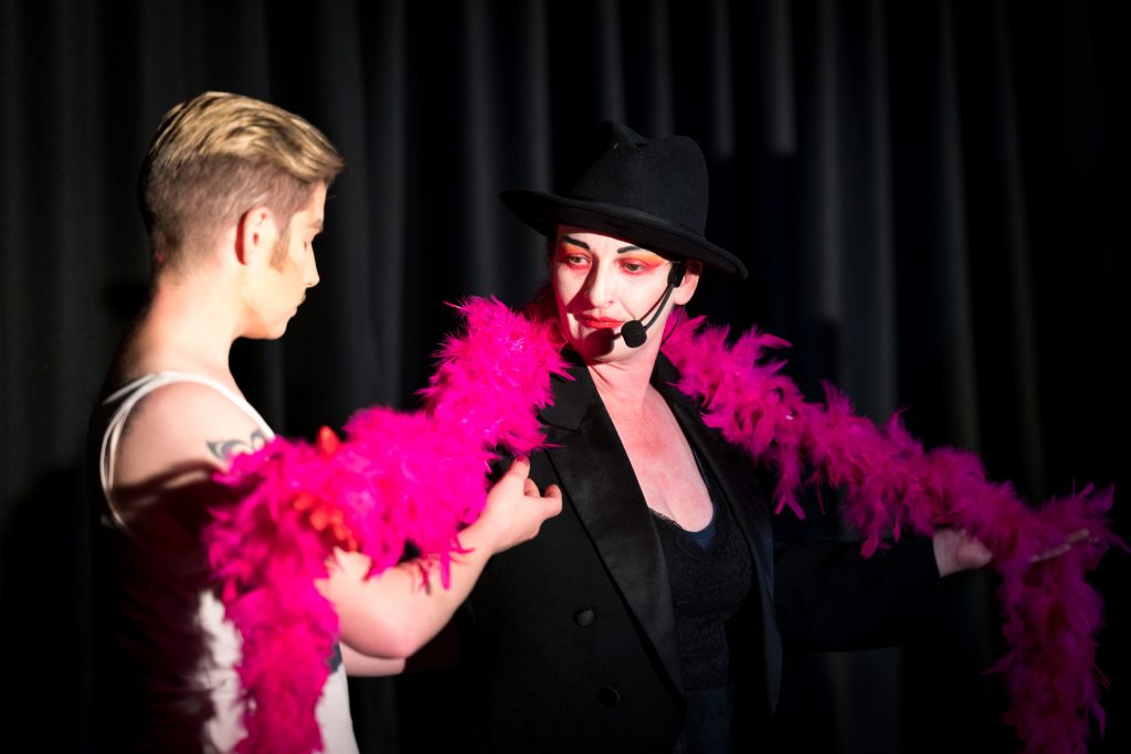 Two drag performers on stage. Right, a white woman with black bowler hat, a long pink feather boa and black suit jacket. The performer on the left is reaching up to the boa.