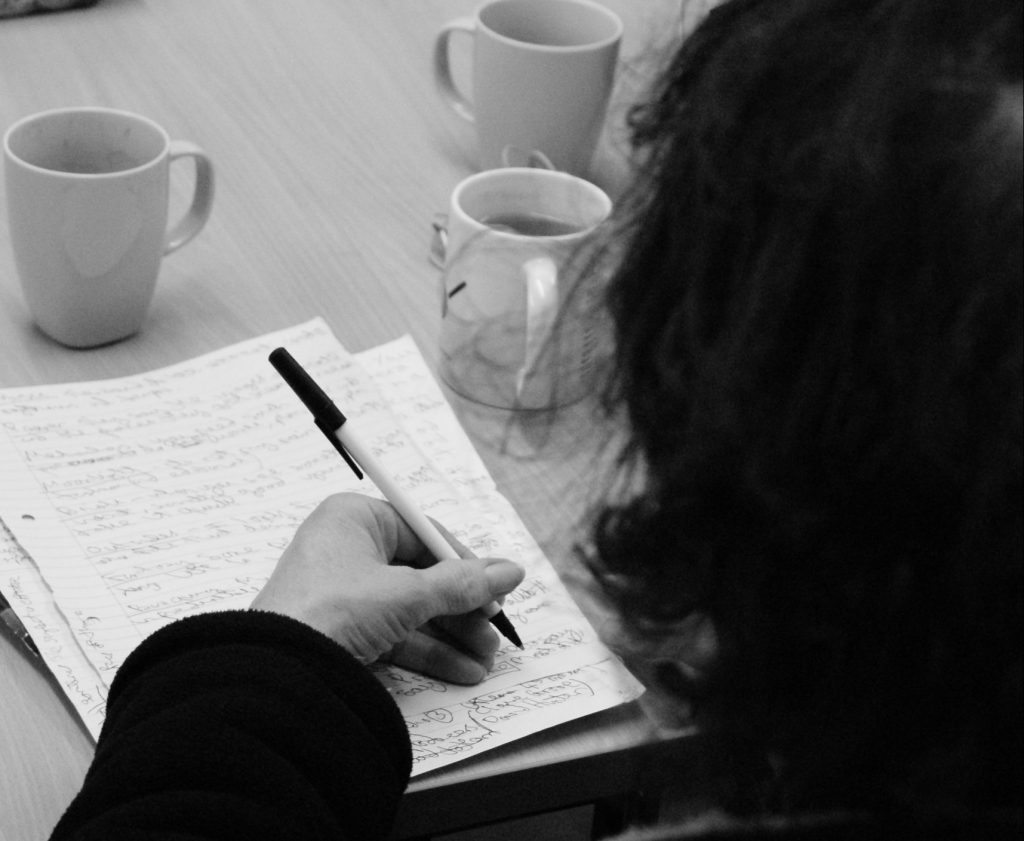 A close crop of someone writing in a notebook at a table, taken from the perspective of someone standing behind their shoulder.