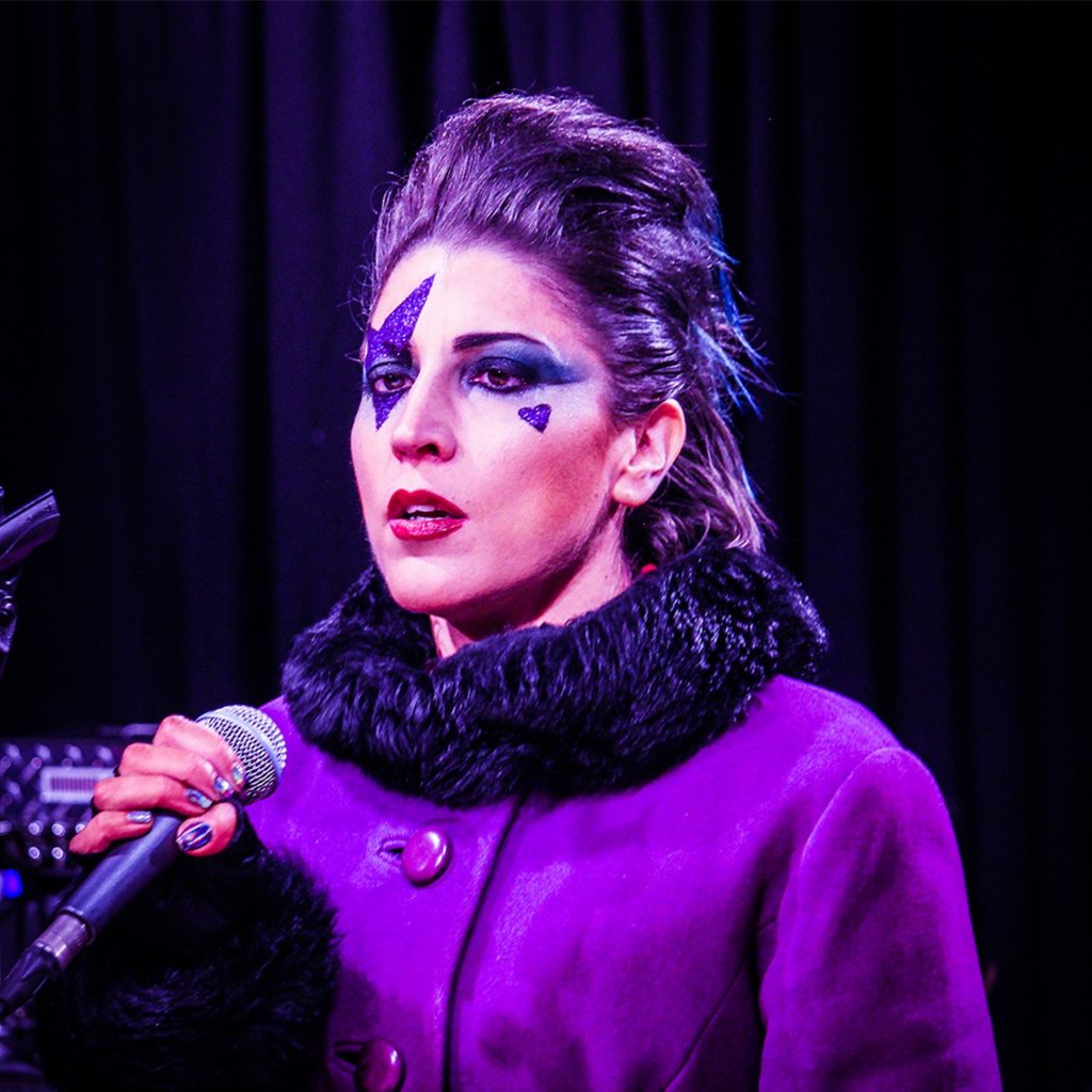 A close up of a white woman with electric blue lightning makeup on her right eye and smoky blue eyeshadow on the other. She is wearing a purple coat with fur collar and grasping a microphone.