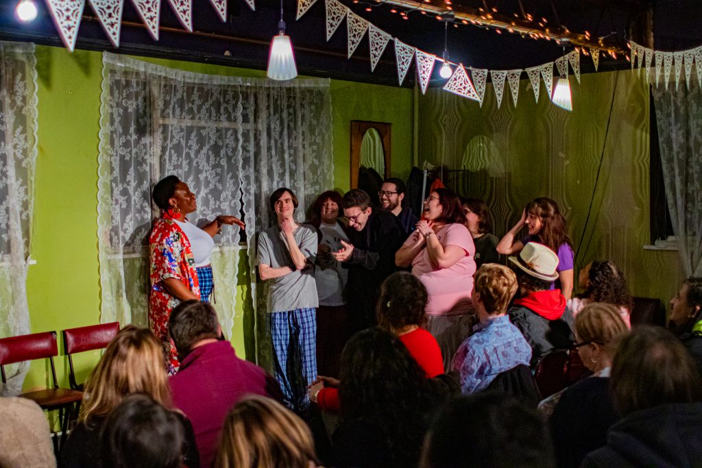 A black woman conversing to a group of seven others who are laughing at what she is saying.