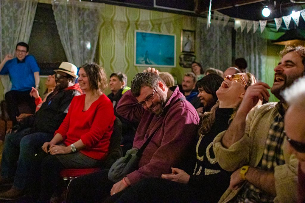 The audience smiling. Centre, a man is itching his head in a sheepish but joyous manner