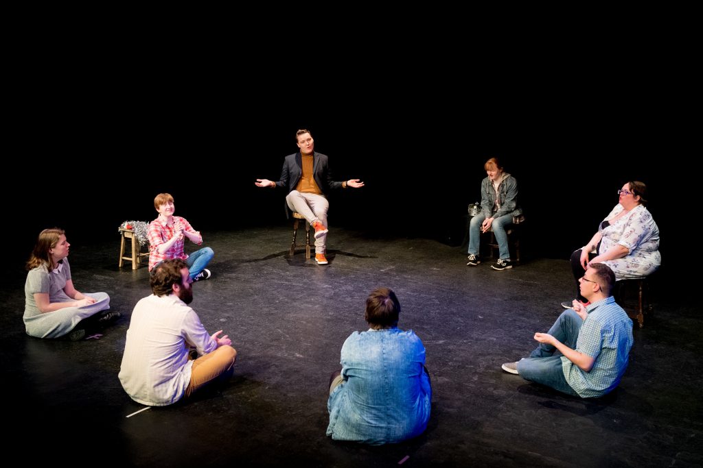 A group of eight workshop participants sitting in a circle on stage in a black box theatre. A man is sitting with legs crossed in the back with his hands in a 'what if' pose while the others sit crosslegged or on chairs with their attention to him