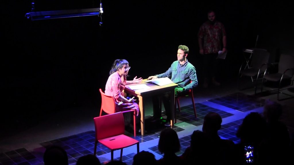 An Asian woman and a young white man sit on opposite ends of a table on stage. The woman is exclaiming to the audience, while the man looks at her.