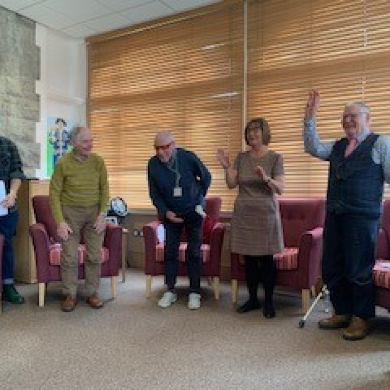 Six participants standing in a half semi circle midway through a storytelling session, an older man on the left has his hands up in dance, a woman next to him is clapping, everyone is smiling and cheery