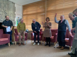 Six participants standing in a half semi circle midway through a storytelling session, an older man on the left has his hands up in dance, a woman next to him is clapping, everyone is smiling and cheery