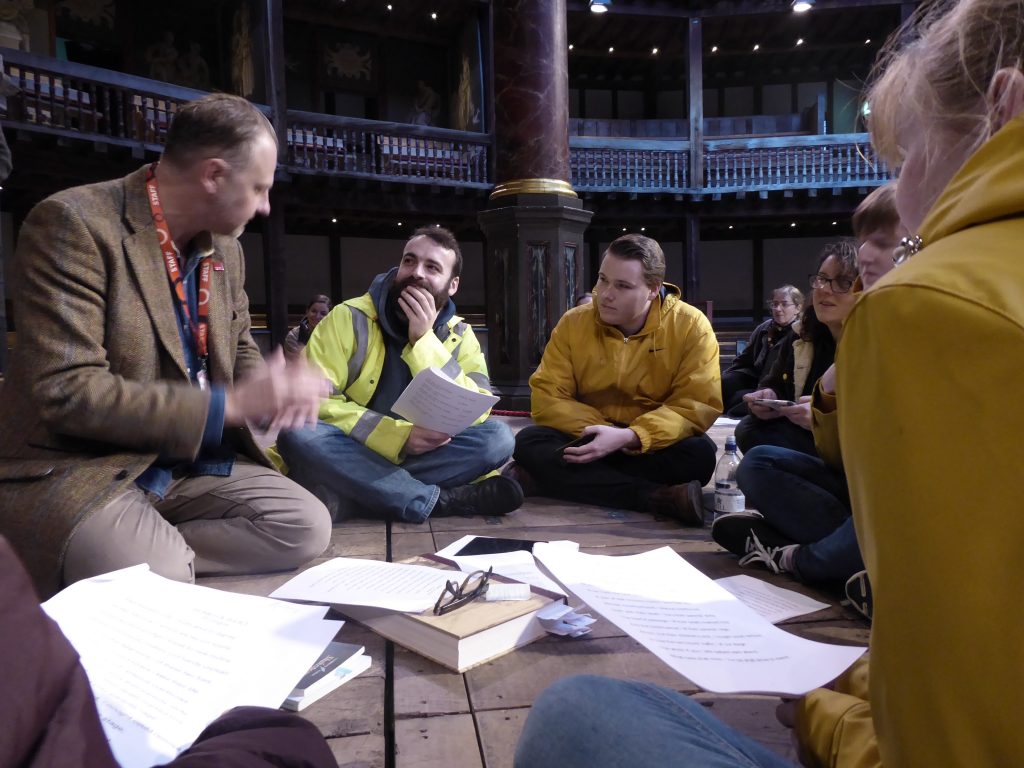 A medium shot of a group of people sitting in a circle on an antique stage looking at scripts