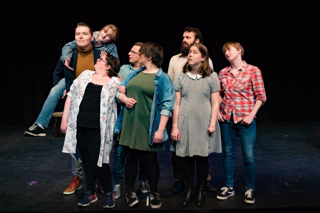 A group of visually impaired and blind actors standing on stage, their attention directed to one of them being piggybacked.