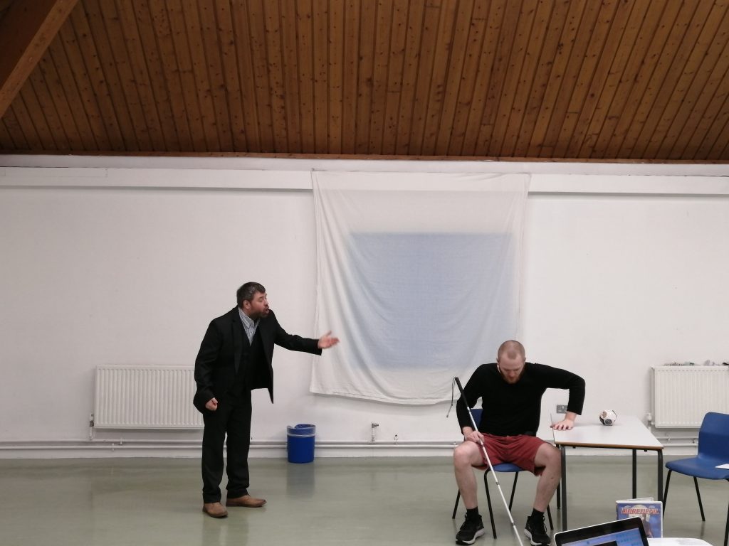 A white man in a suit exclaims to a younger white man sitting down who is seemingly lost in thought