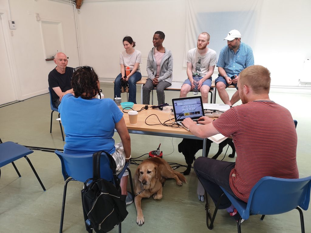 Five actors and the stage manager sit and listen while the director feeds back. A guide dog lays at the director's feet.