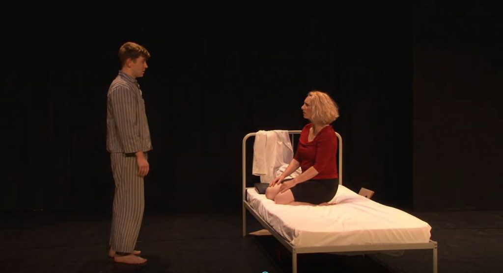 A white man dressed in striped pajamas stares solemnly towards a white woman dressed in a red top and black skirt kneeling on a white hospital bed.