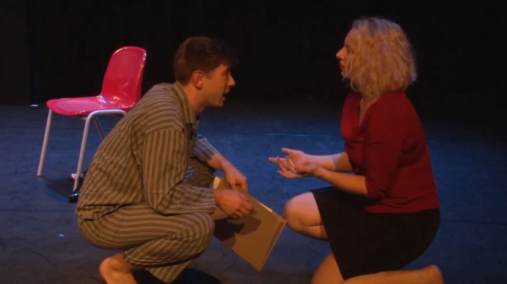 A white man in striped pajamas kneels on the floor talking to a white woman dressed in red top and black skirt, also kneeling.