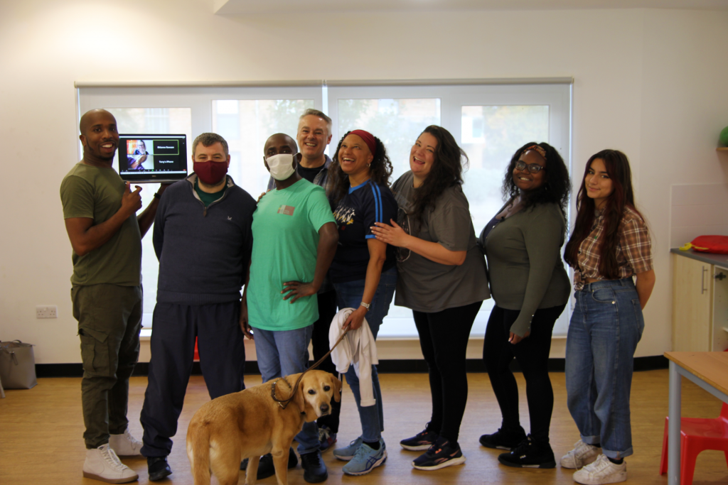 A group photo of the Super Power Panto R&D team standing next to each other in the rehearsal room, and a guide dog in the middle.