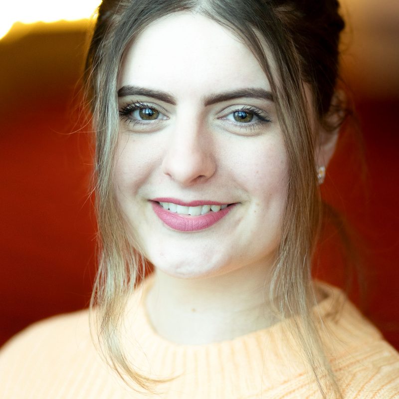 Headshot of a white brunette woman, smiling and wearing an pale orange jumper.