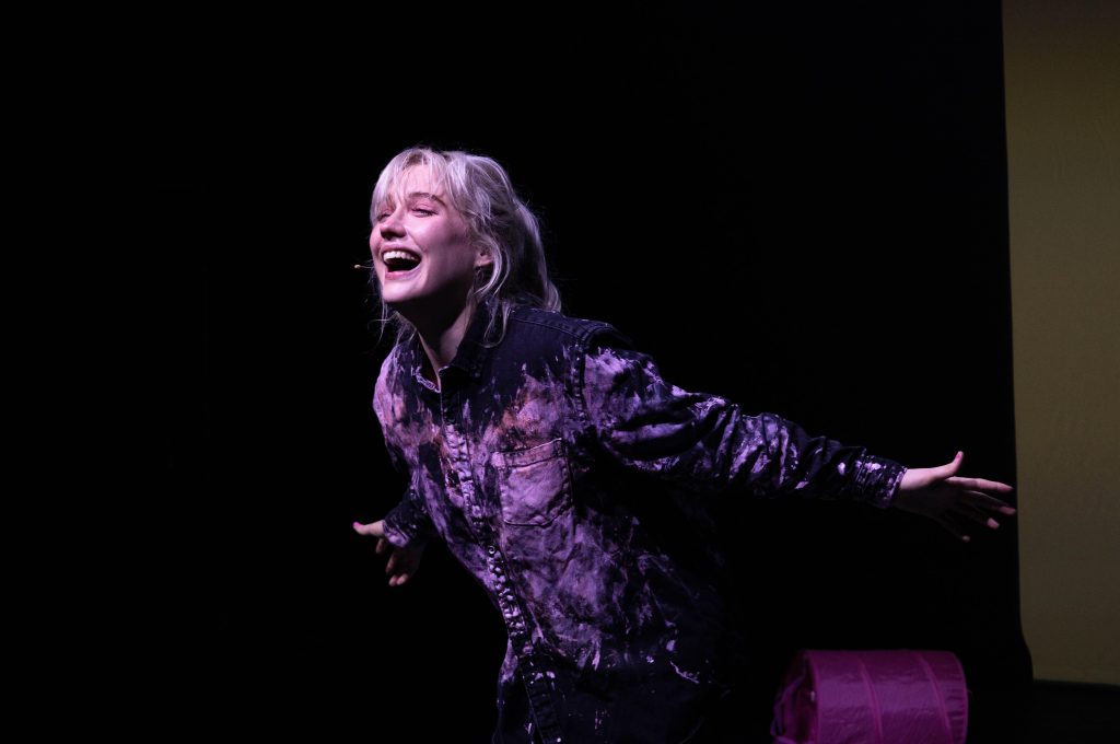 production shot of Alice, a white blonde woman in her mid twenties, performing in Past Life. She is stood leaning forward with arms outstretched and laughing, with soft lighting on her face and is wearing a dark pink tie-dye shirt. 