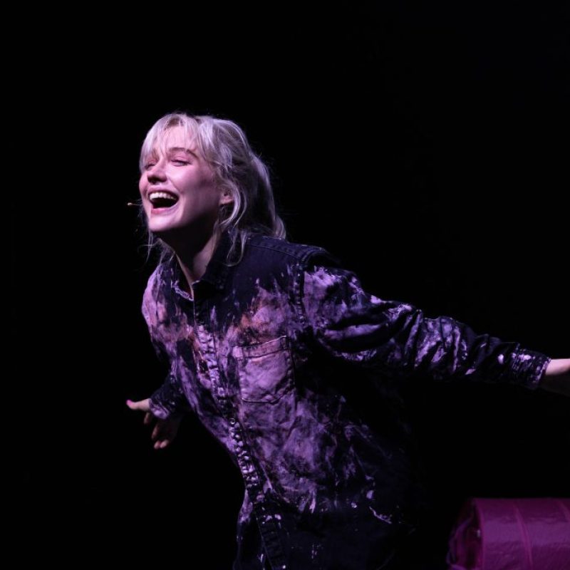 production shot of Alice, a white blonde woman in her mid twenties, performing in Past Life. She is stood leaning forward with arms outstretched and laughing, with soft lighting on her face and is wearing a dark pink tie-dye shirt.
