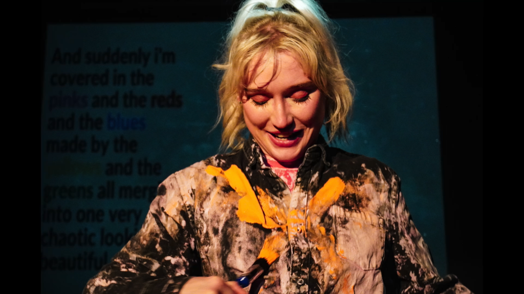 Alice is wearing a black shirt covered in streaks of white paint, and is using a brush to daub an X across her chest in yellow paint.