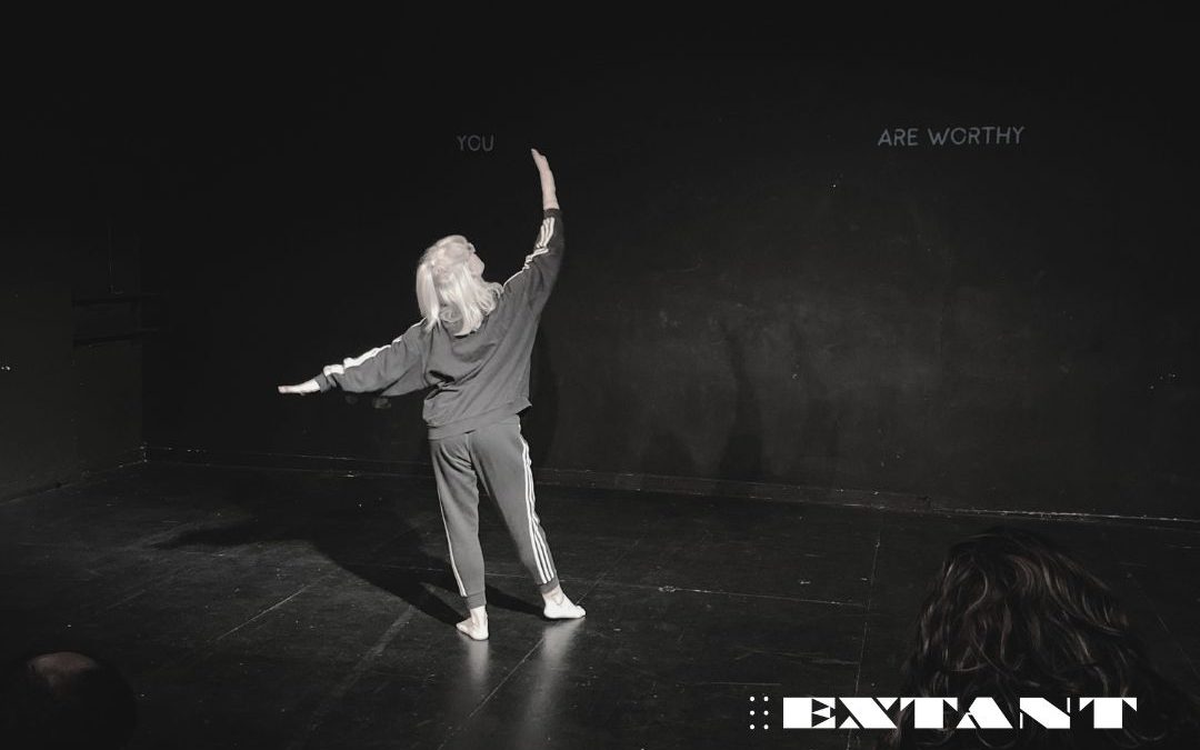 A photograph of Alice Christina-Corrigan, wearing matching black tracksuit trousers and top. She faces away from the camera towards the back of the stage, her right arm lifted to the ceiling and her left reached out sideways. On the back wall there is a projection of the words 'You are worthy' which is separated so the word 'You' is on the far left and the words 'are worthy' are on the far right. Edited onto the photo is the Extant logo in the bottom right corner of the image.