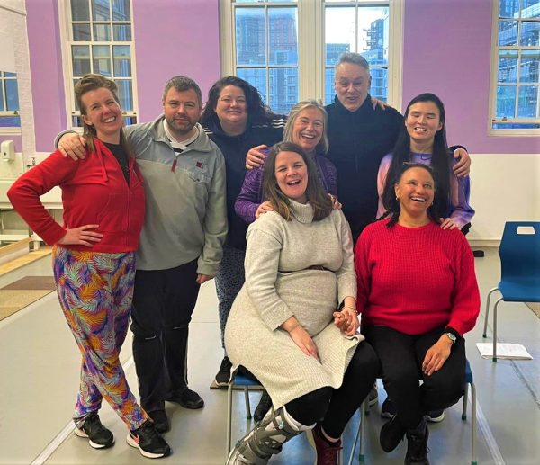 A group photo of the Super Power Panto team - they are eight adults of a range of ages and ethinicities with their arms around each other smiling towards the camera in a rehearsal room.