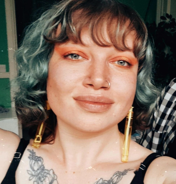 Close of shot of Rhianna, a white woman in her mid 20s with blue eyes and brown and dyed blue hair, a nose ring and long yellow earrings looking straight into the camera.