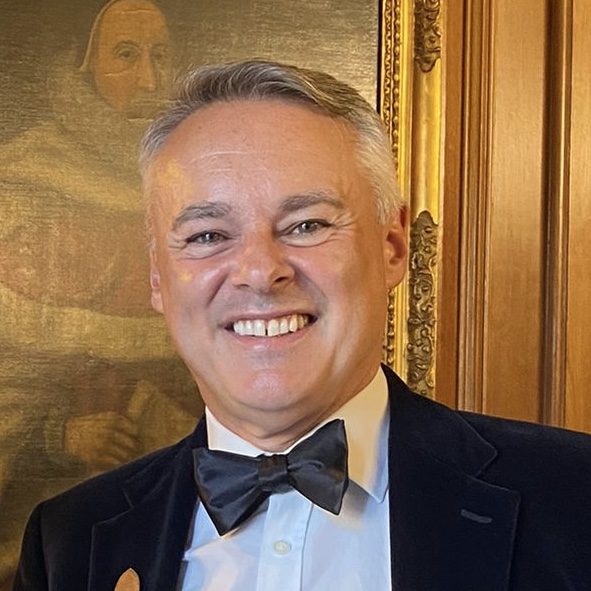 Headshot of Chris, a middle aged white man with greying hair smiling in front of a painting in a bow tie, white shirt and smart jacket.