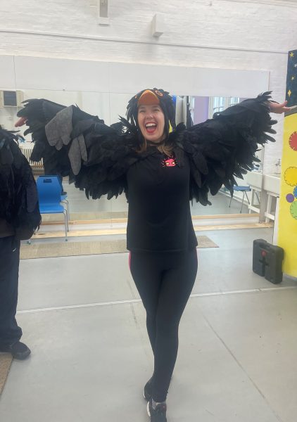 Angela, a white woman with brown hair, wears a black bird costume with feathery wings and a hat with an orange beak. She raises her arms either side of her as if flying and smiles towards the camera. 