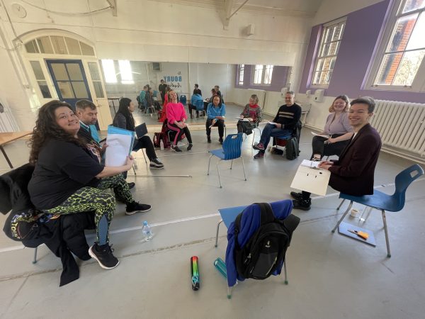 A group of people of varying ages of ethnicities sit on blue plastic chairs in a rehearsal space holding scripts. It is the first read through of Super Power Panto.