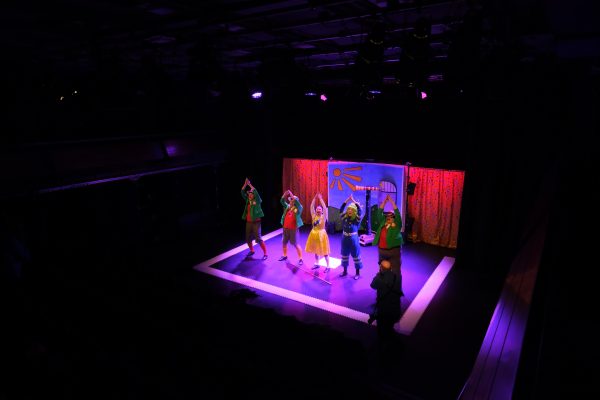 A shot from high up at the back of the auditorium of the cast of Super Power Panto onstage in the middle of the show's final number. Three of them wear blazers, boater hats and long socks, one a yellow dress and one a blue boiler suit with a bright green wig. They all have their hands together raised above their heads.