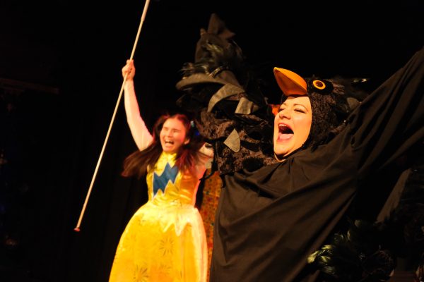 In the foreground mixed heritage actor Chloe wears a blackbird outfit with feathery black wings and a headpiece with an orange beak and big eyes. She raises her arms at her sides as though in flight and has her mouth open in song. In The background, Jasmin, a chinese actor wearing a yellow dress with a blue lightning bolt across the chest raises her arms too, in her right hand she holds a white cane.
