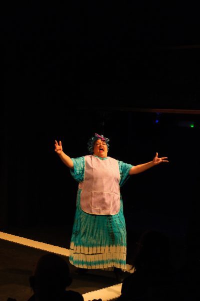 Mixed-heritage actor Chloe dressed as mum in a long patterned blue dress, apron and blue bonnet stands alone on a corner of the stage with her hands outstretched in an operatic manner.