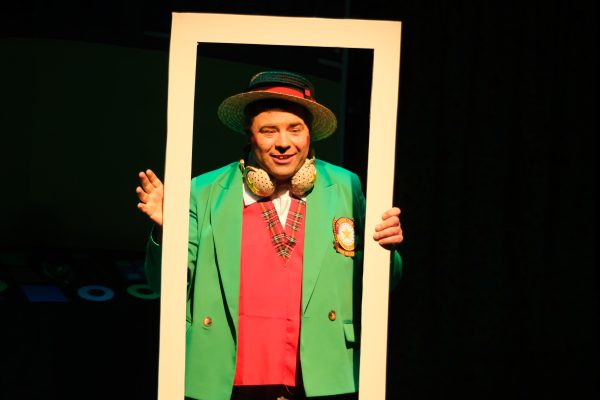 White actor Steve stands in a spotlight in the middle of a tall white rectangular frame, smiling. He wears a boater hat, green blazer with a red waistcoat underneath and a pair of golden headphones around his neck.